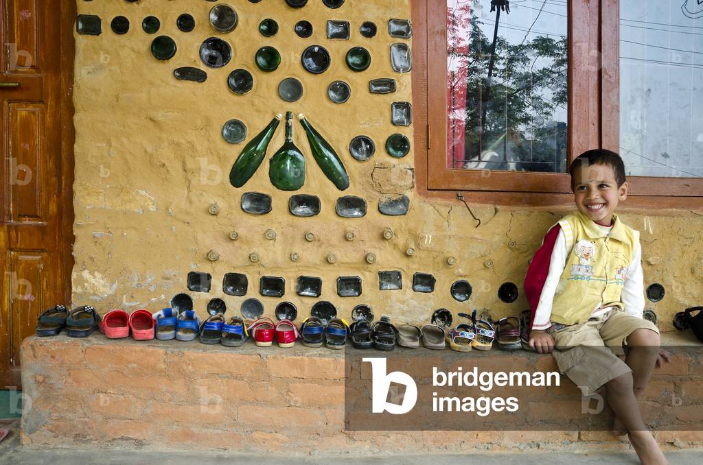 Nepali boy sitting outside bottle house, lined up pairs of sandals of children inside nursery/school building, Kahare, Chalnakhel, Kathmandu Valley, Nepal (photo)