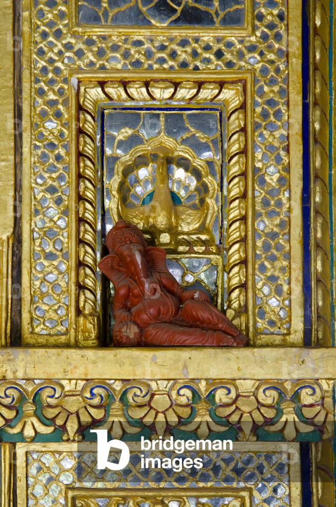 Hinduism: Small reclining Ganesha (elephant-headed deity) statue on ornate gilded and mirrored wall ledge in old Rama (avatar of Vishnu) temple(sharing premises with the Nirvana cafe/restaurant)The Old Blue City, Jodhpur, Rajasthan, India