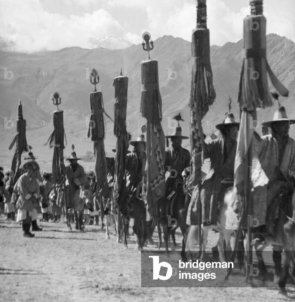 Horsemen in ceremonial dress and wearing conical hats with brims carrying banners and standards surmounted by tridents, Doguthang, Rikya, Tibet, 8th October 1939 (b/w photo)