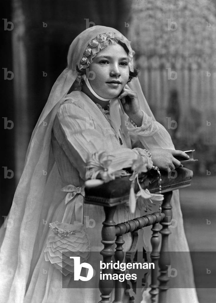 Girl making her first communion c. 1955, France: she's kneeling on a prie-dieu, she's wearing a white dress with purse and veil, she's holding a missal