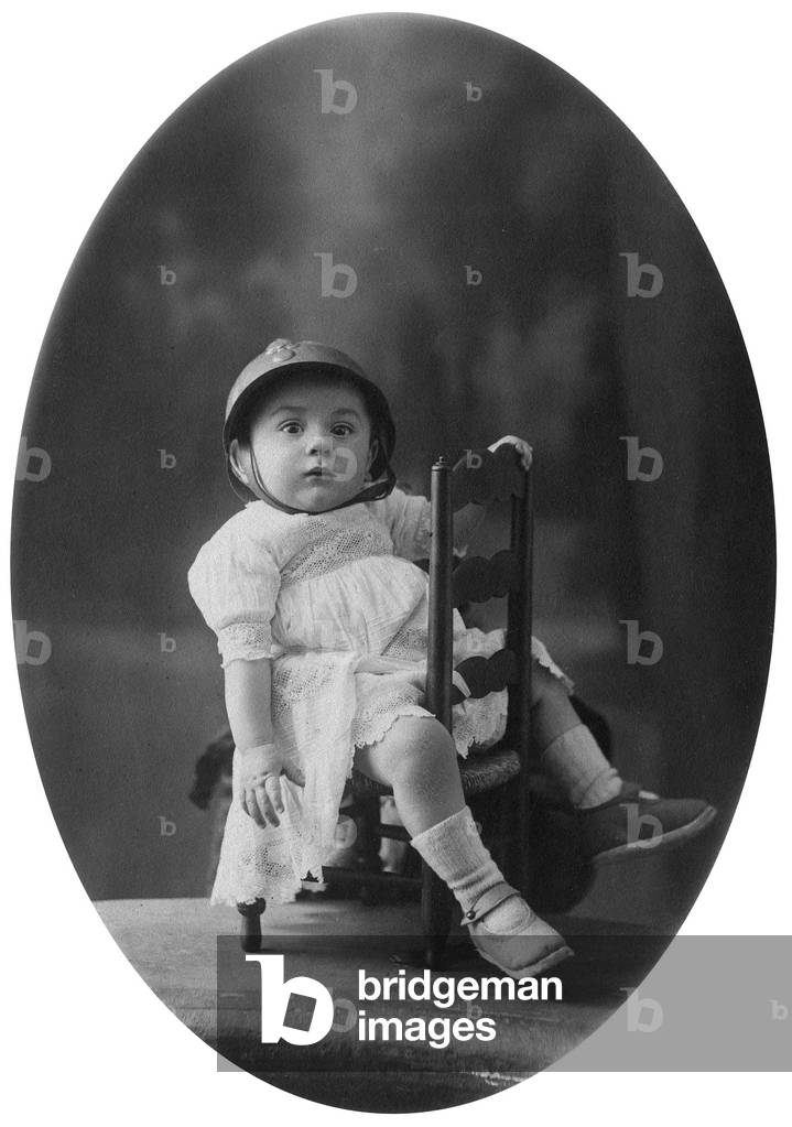 Baby on a chair with an helmet, c. 1920 (photo)