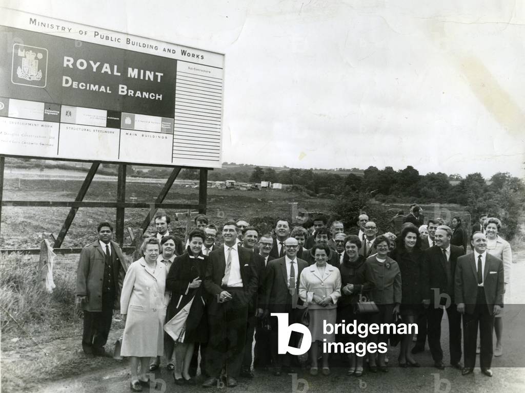A group of Mint employees from London gather by the site sign of the new Royal Mint at Llantrisant, 1967 (b/w photo)