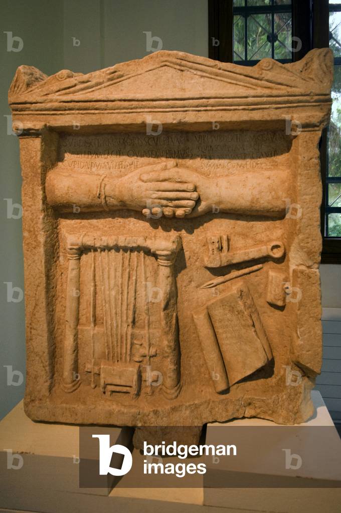 Funerary stele with a dexiosis or handclasp.Nabilium (string instrument), a key, an inkwell, and an open book roll with Latin verse. 1st century AD. Dion, Greece,