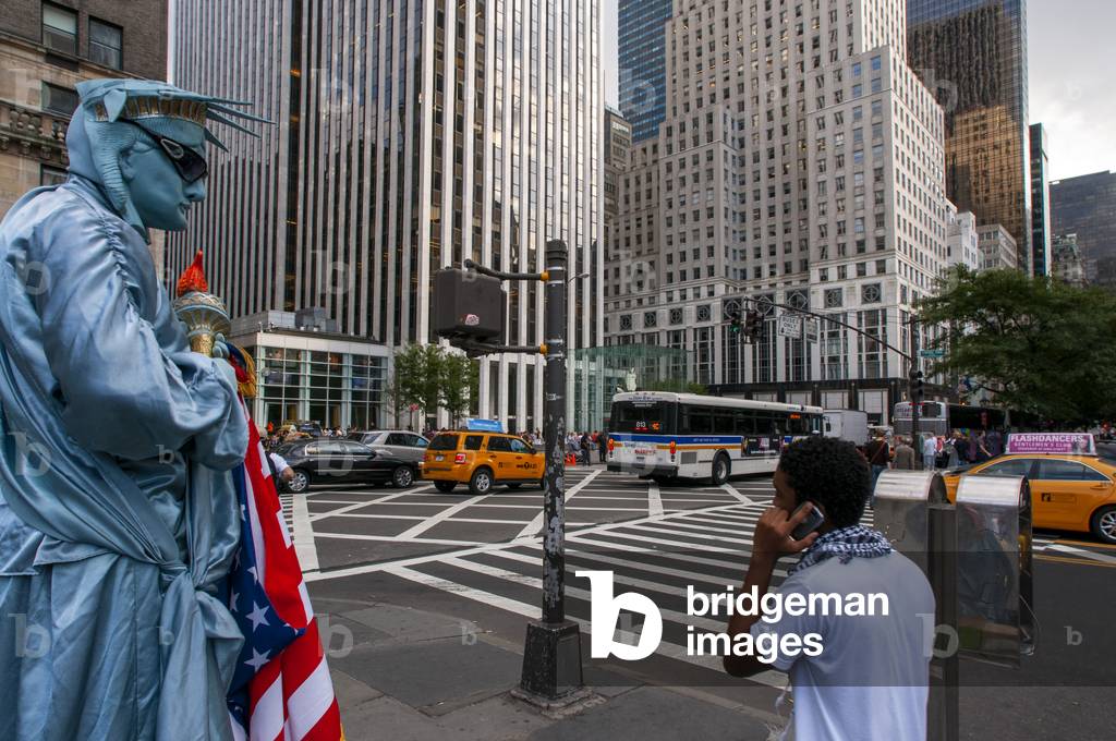 A human statue dressed in Statue of Liberty with tourists waiting to be photographed at the exit of Central Park plaza overlooking the Grand Army Plaza, New York City, USA (photo)
