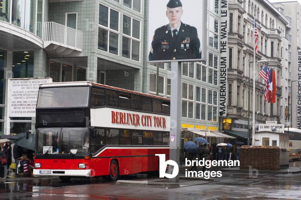 Berliner city tour bus sightseeing in front of Checkpoint Charlie US Army checkpoint and guardhouse reconstruction at the former crossing point between East and West Berlin in Berlin, Germany (photo)
