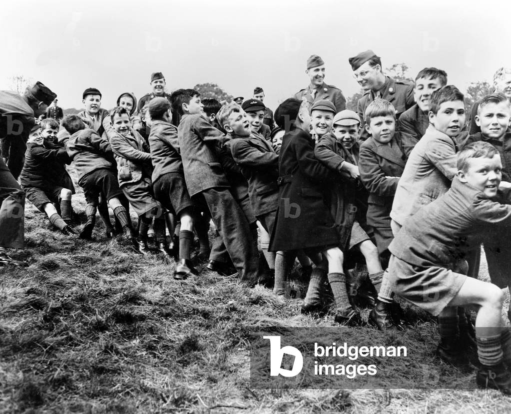 Village children in a game of Tug-of-War being cheered on by US airmen who were stationed near their village during World War II (b/w photo)