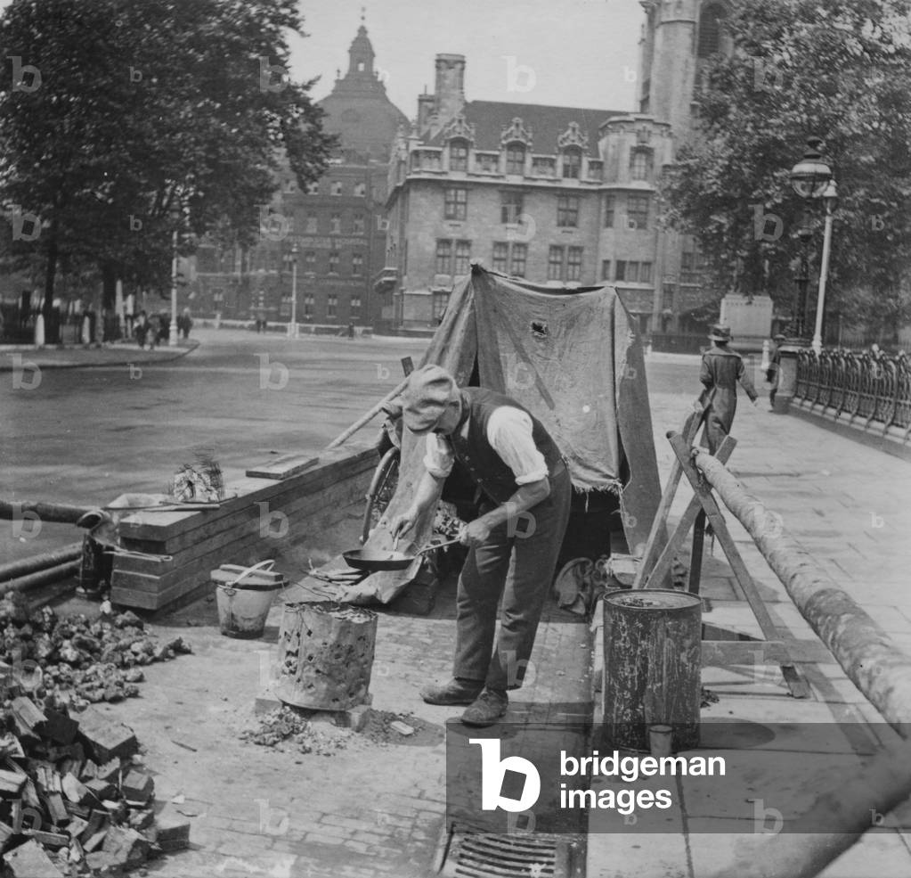 Construction worker enjoying a break, cooking over a makeshift stove by the side of the pavement, London, c.1910-20 (b/w photo)