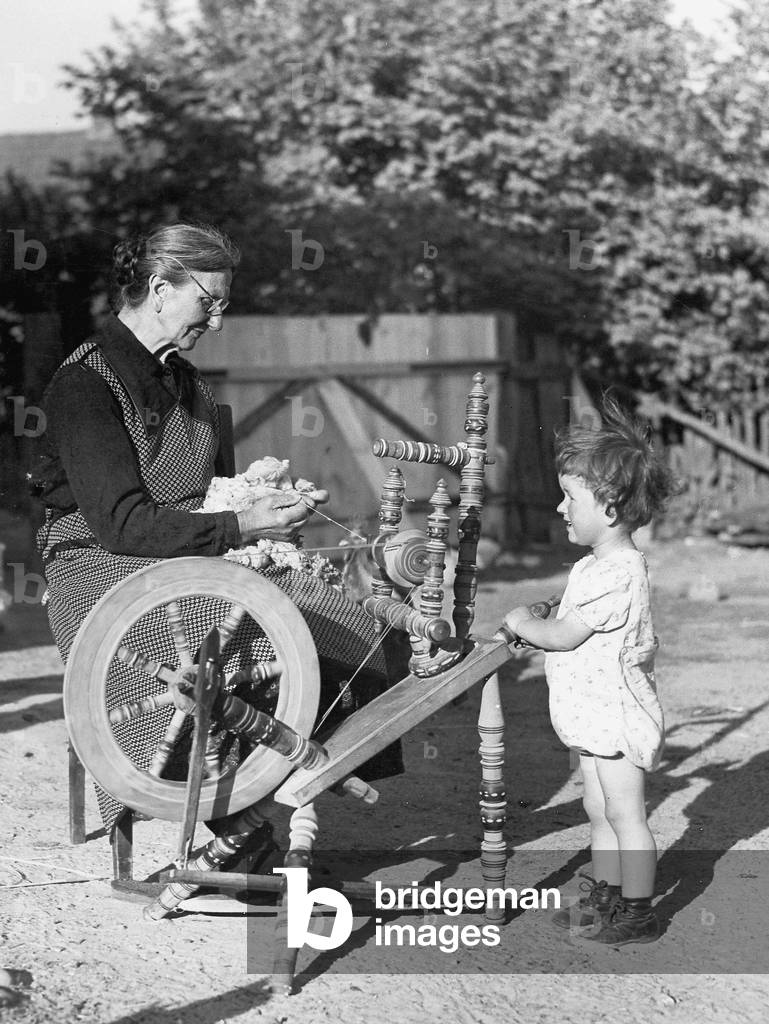 Eine ältere Frau spinnt Wolle mit ihrem Enkelkind auf (s/w Foto)
