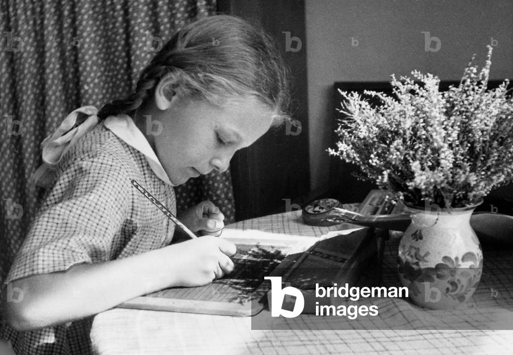 A girl seated at a table does her homework on a slate, 1941