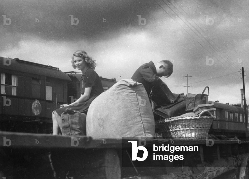 Boy and girl sitting with their belongings on a platform, waiting for their train, 1945 (b/w photo)