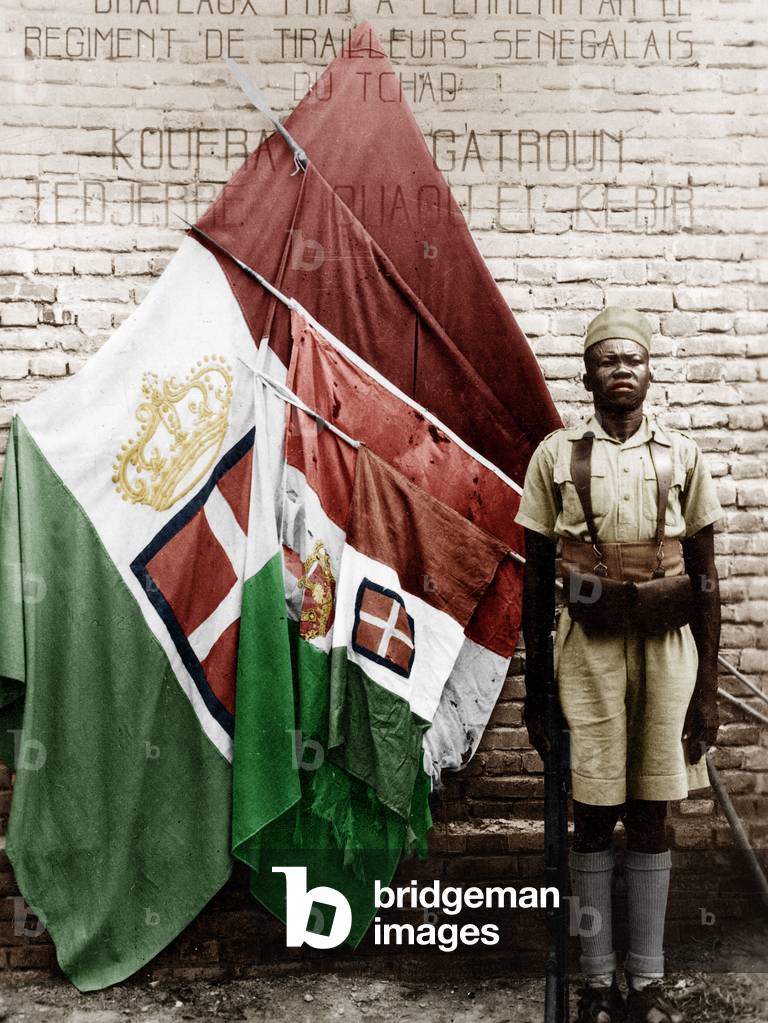 Soldier of the Regiment of the Senegalese Riflemen of Chad in front of the flags taken to the Italians during the countryside of Libya and of Fezzari, during the Second World War, 1939-1945 colourized document