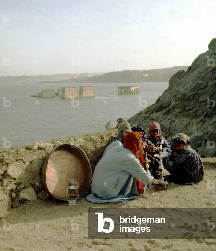 Young boys smoking the Narguile. In the distance, the temple of Isis in Philae before the move by UNESCO, Aswan, 1968.