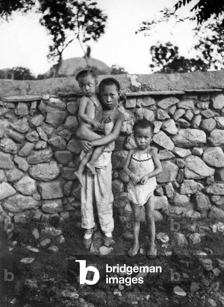 China: c. 1925 - Three children standing in front of a stone wall in a small country village in rural China.