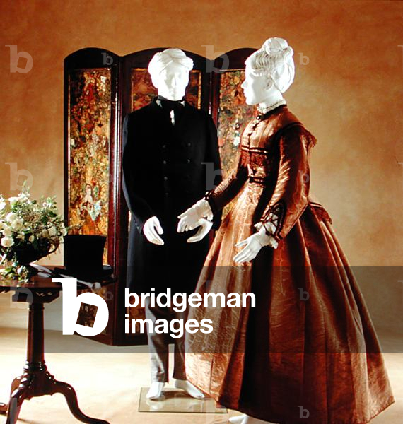 Una signora in corpetto marrone moire e gonna con nastro di velluto e pizzo, c.1869, e un uomo in un abito ner