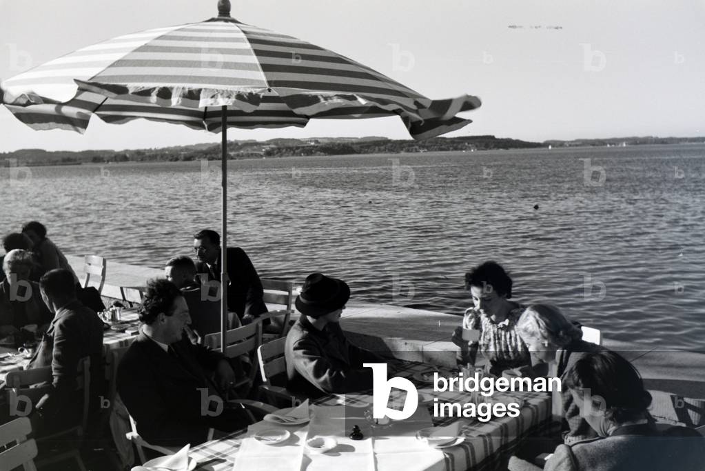 Guests of a café at the Chiemsee sitting under sun shades on the terrace, Germany 1930s (b/w photo)