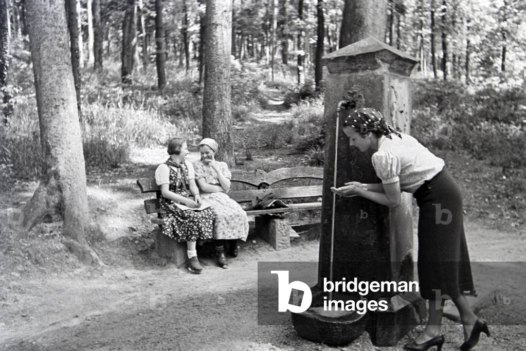 A young woman is tasting the drinking water of the Eberbrunnen on a hiking path near Baden-Baden in the Northern Black Forest, Germany 1930s (b/w photo)