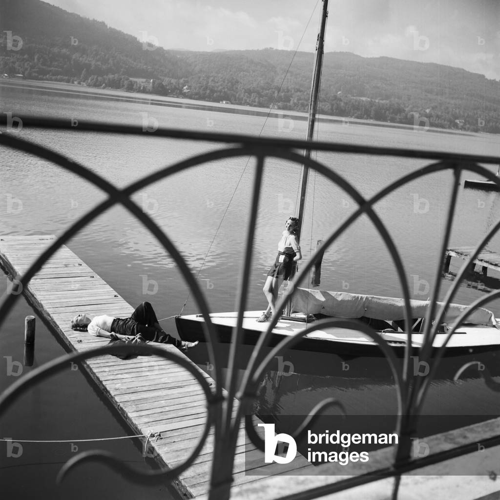 Two young woman tanning on the shore of a lake, one on a boardwalk, the other on a sailing boat, Germany 1930s (b/w photo)