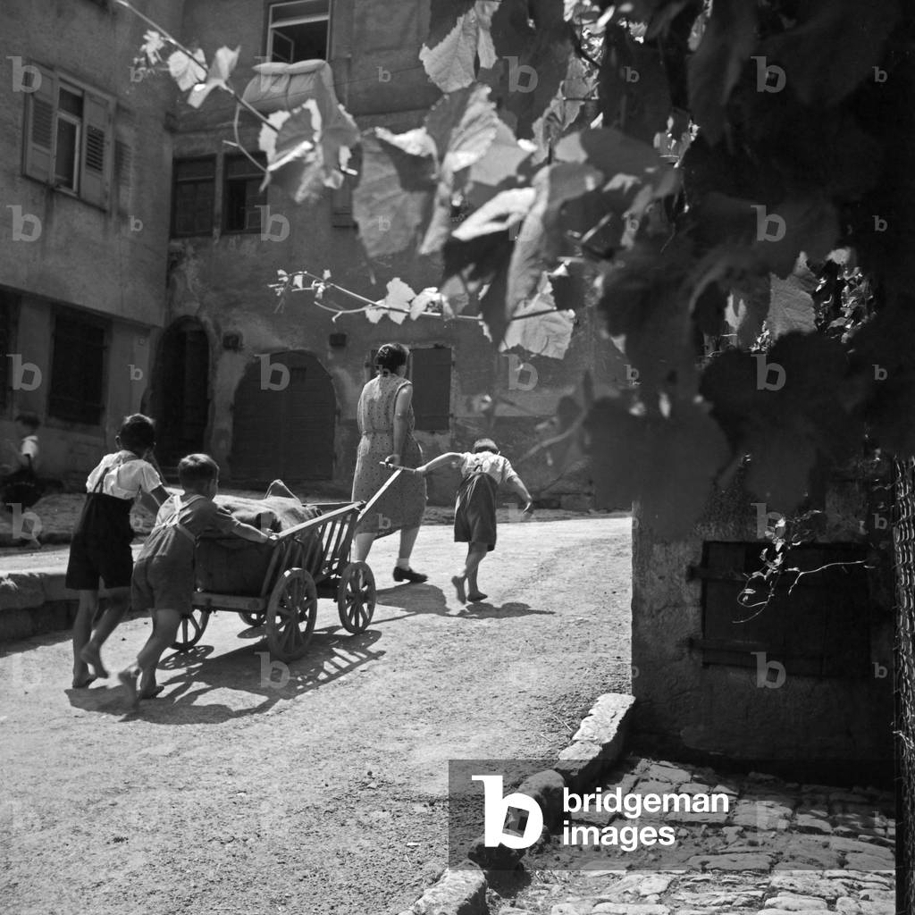 A woman and three children pulling a push cart through the lanes of Rothenburg ob der Tauber, Germany 1930s (b/w photo)