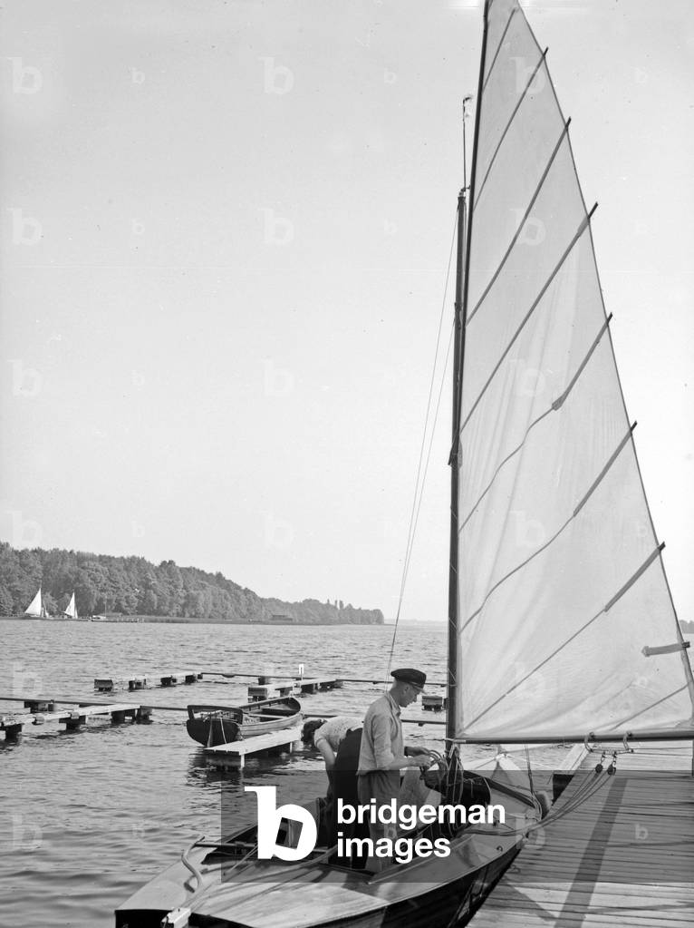 A sailing boat anchoring at a boarwalk at the shore of a lake, Germany 1930s (b/w photo)