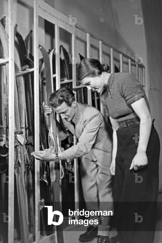A skiing teacher helping a femal beginner to chose the right skis, Germany 1930s (b/w photo)