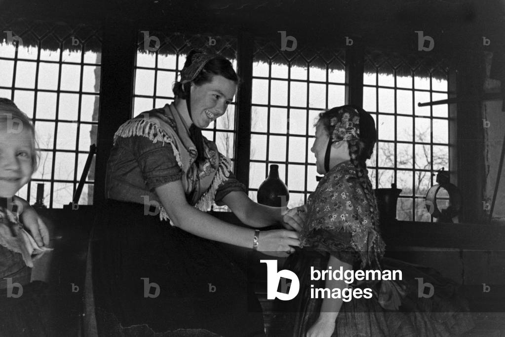 A woman helping a little girl to wear traditional costume of Ammerland near Bad Zwischenahn at the Oldenburg area, Germany 1930s (b/w photo)