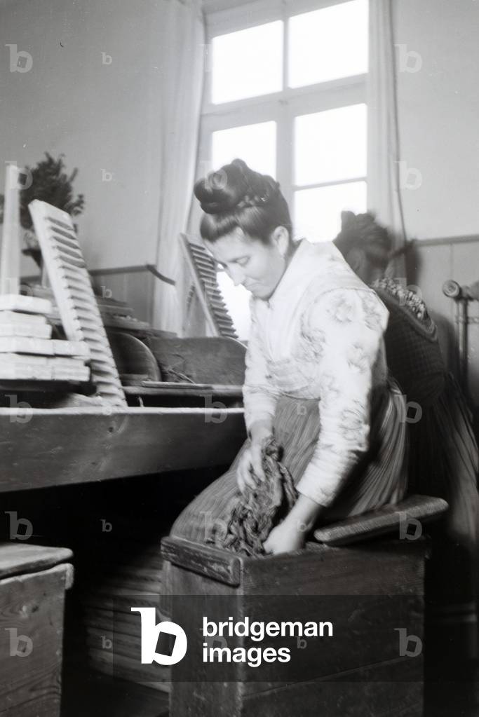 A female cigar roller wearing a Marburg garb, costume is taking tabacco leaves out of a trunk that serves her as a seat at the same time, near Marburg, Germany 1930s (b/w photo)