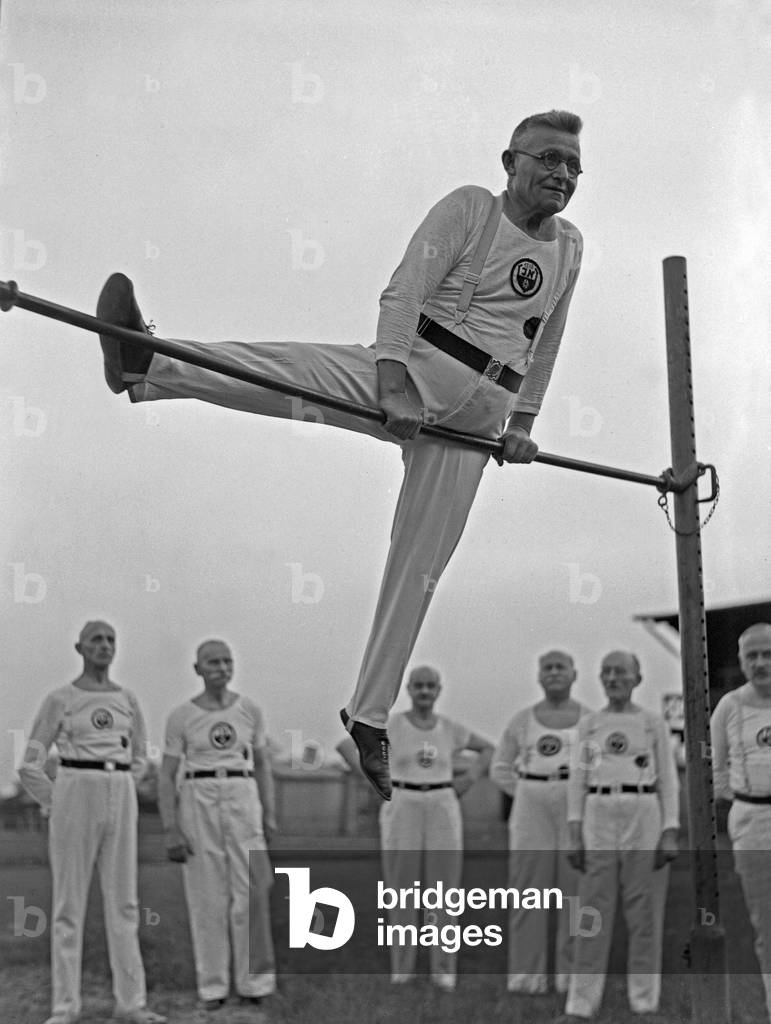 A member of the senior department of a gymnastic club performing at a high bar on a sporting ground, Germany 1930s (b/w photo)