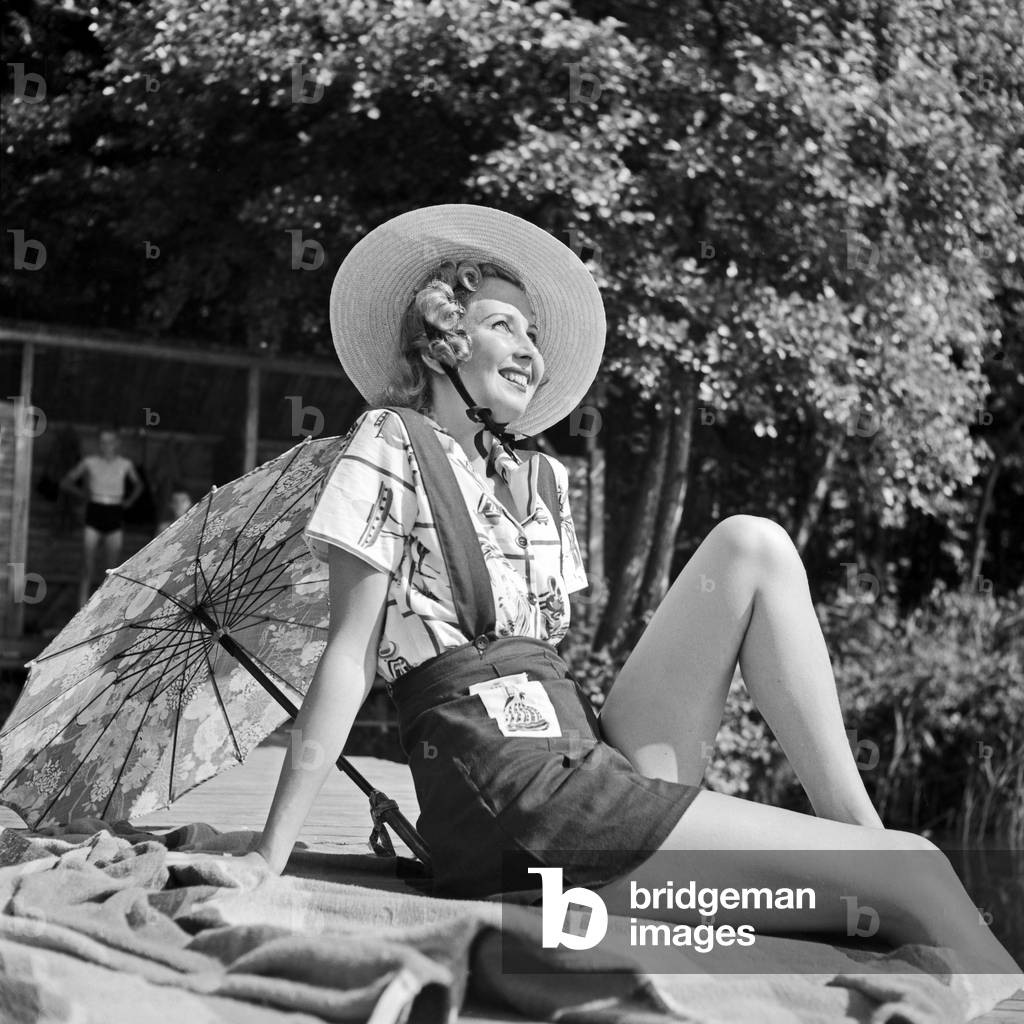 A young woman in beach dress sitting on a boardwalk at a lake, Germany 1930s (b/w photo)