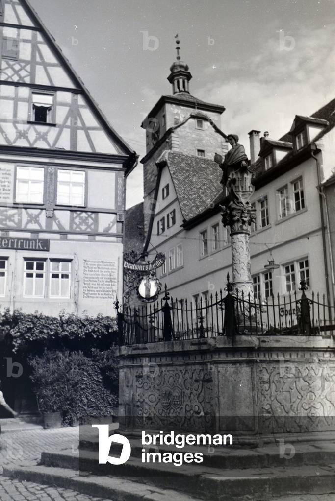 A fountain with a litle statue in Rothenburg ob der Tauber, with the gothic part and the high towerin the background, Germany 1930s (b/w photo)