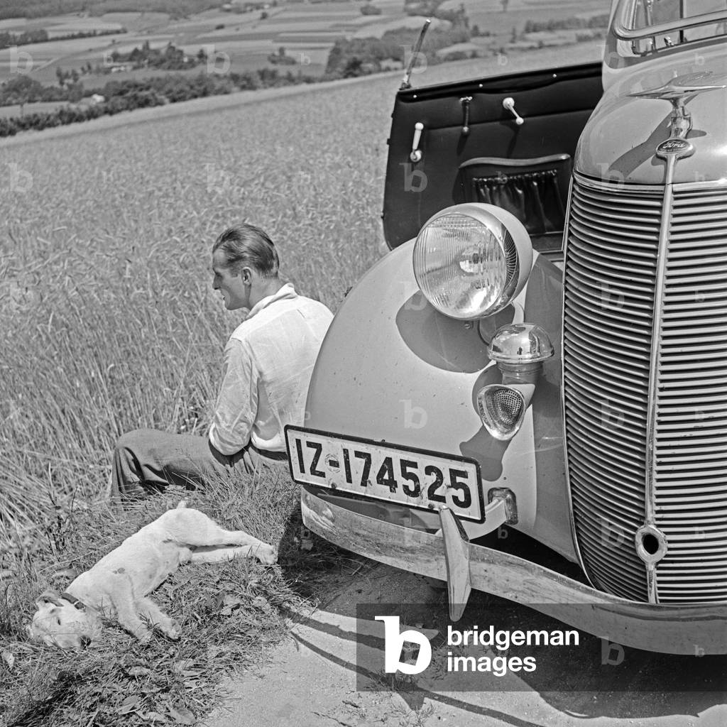 A man and a fox terrier pup sitting in front of their Ford V8 while resting from a trip, Germany 1930s (b/w photo)