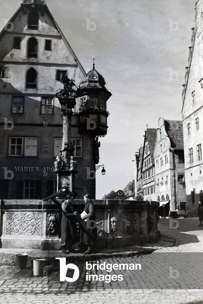 Two women having a chat while the bucktes are filling with water from the fountain placed in front of the impressive town hall in Rothenburg ob der Tauber, Germany 1930s (b/w photo)