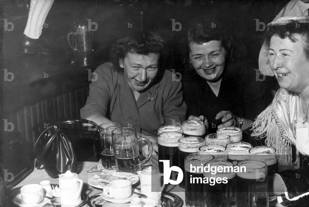 A waitress in a Bavarian style bar serves beer to two very thirsty women, 1950s (b/w photo)