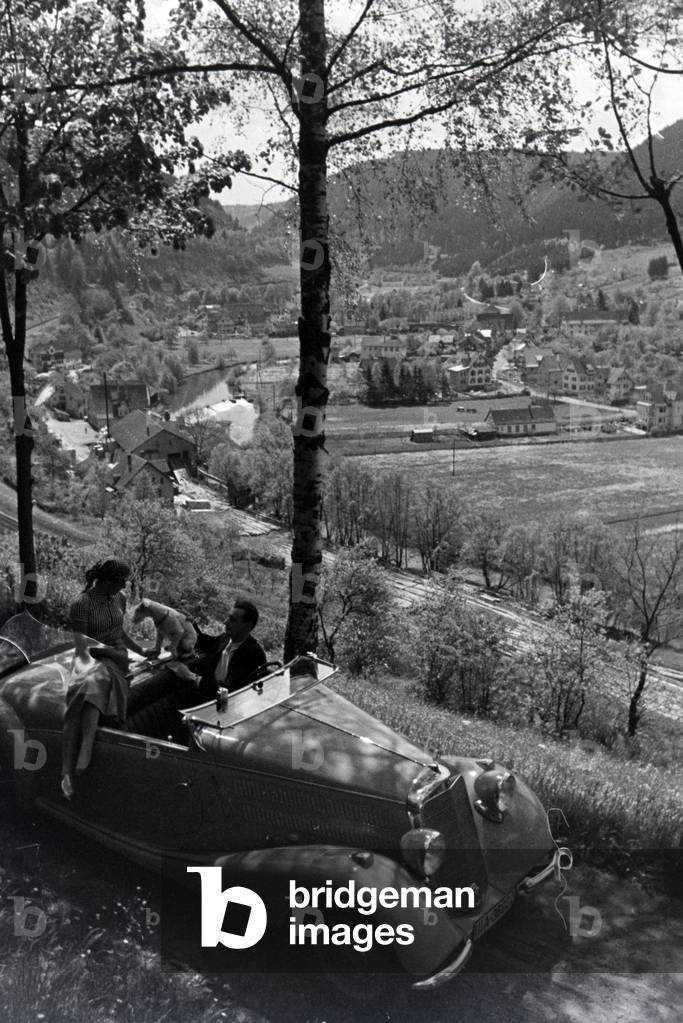 A young couple and their dog driving through Hirsau in the Northern Black Forest in a Mercedes convertible, Germany 1930s (b/w photo)