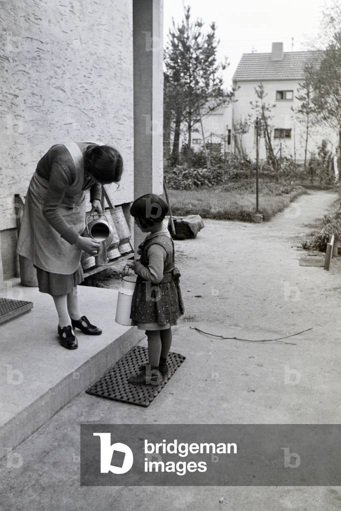 A woman is filling up a mug with milk out of a big milk can for a little girl, zeppelin village near Frankfurt am Main, Germany 1930s (b/w photo)