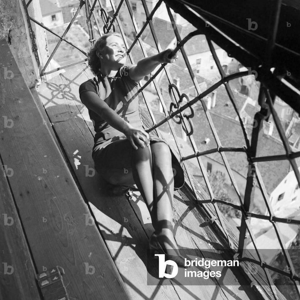 A young woman sitting on the window ledge of a beldry watching down on the city, Germany 1930s (b/w photo)