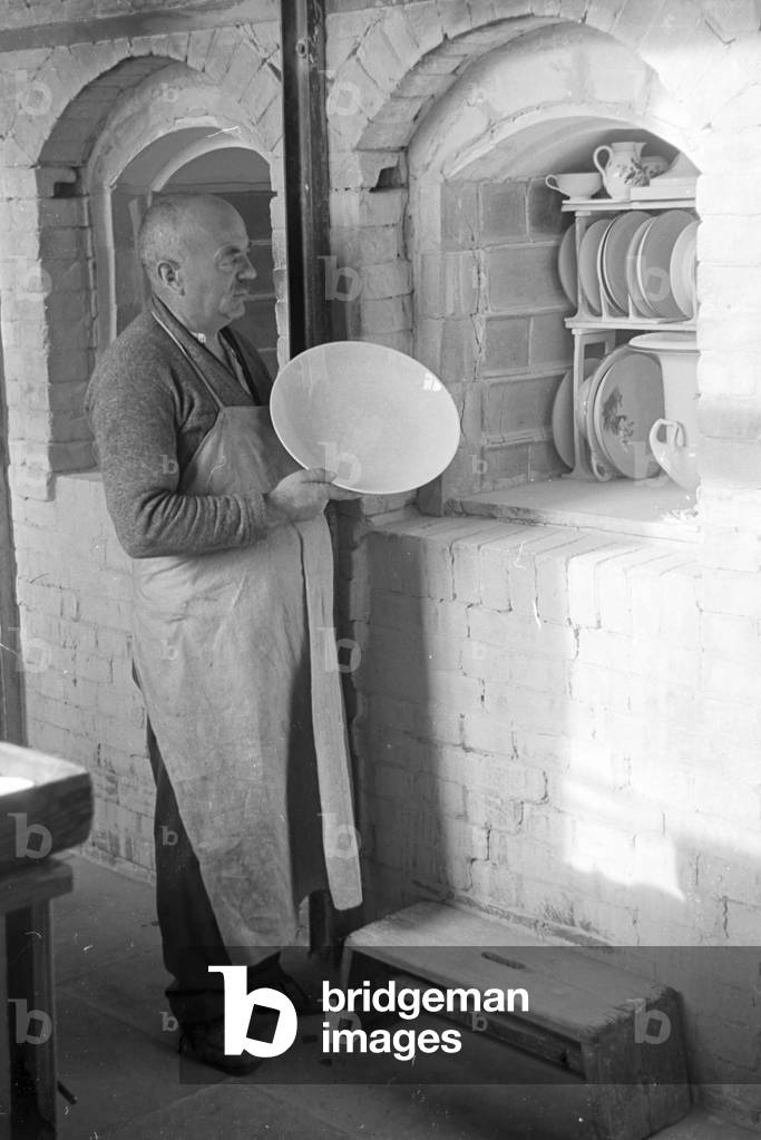 A ceramist of the State Porcelain Manufactory Berlin puts a painted plate inside the kiln for the glost firing, Germany 1930s (b/w photo)