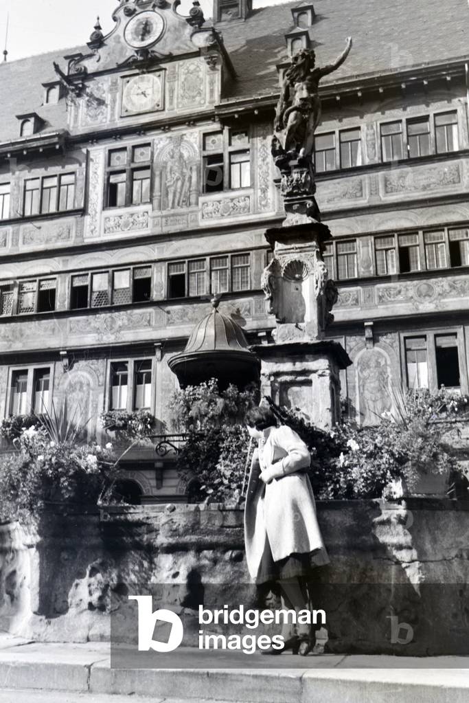 A young woman is posing in front of a fountain at the market square in front of the city hall of Tübingen, Germany 1930s (b/w photo)