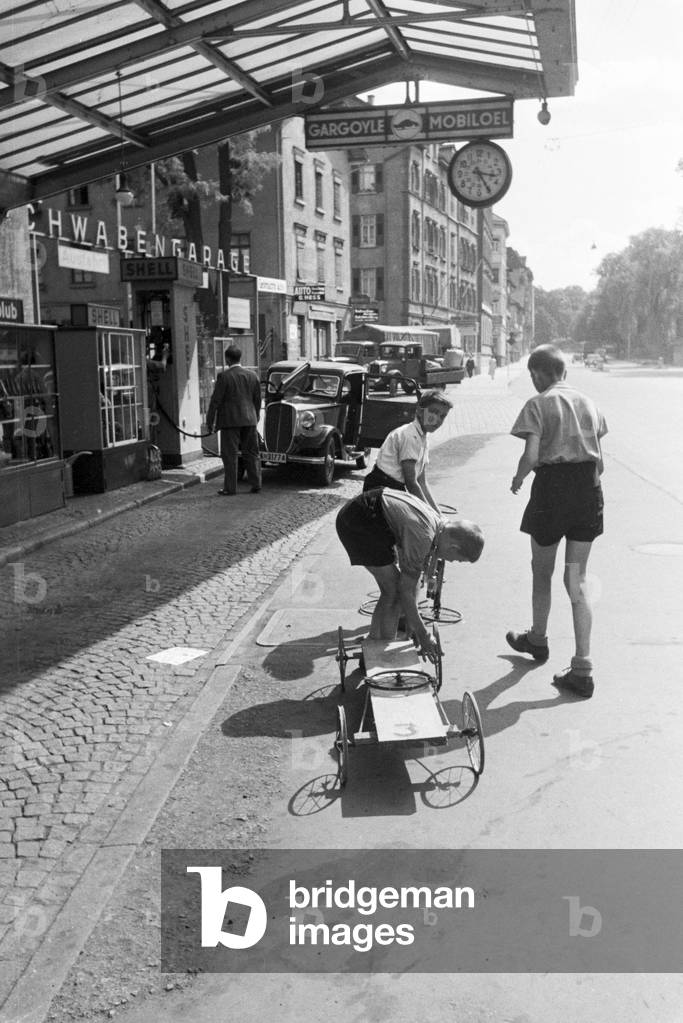 School boys oiling the wheels of their self-built wooden cart at a gas station, Stuttgart, Germany 1930s (b/w photo)