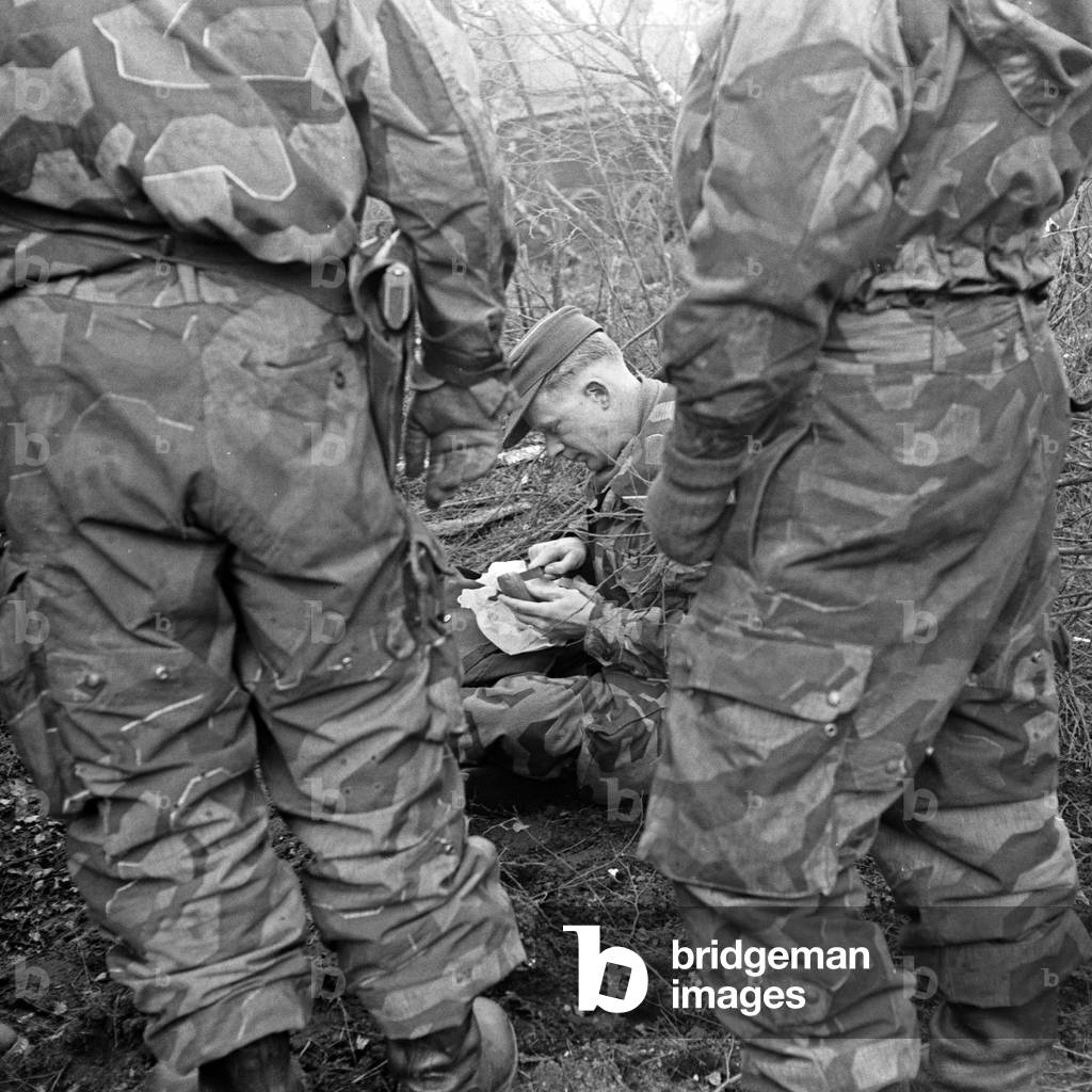 A soldier having a break and eating a sausage at a military exercising ground, Germany 1950s