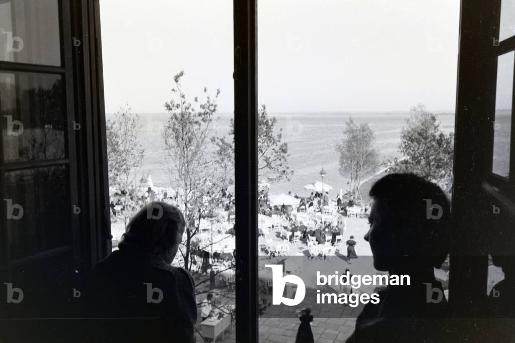 Two young women standing at a window looking down at a café terrace at the Chiemsee, Germany 1930s (b/w photo)