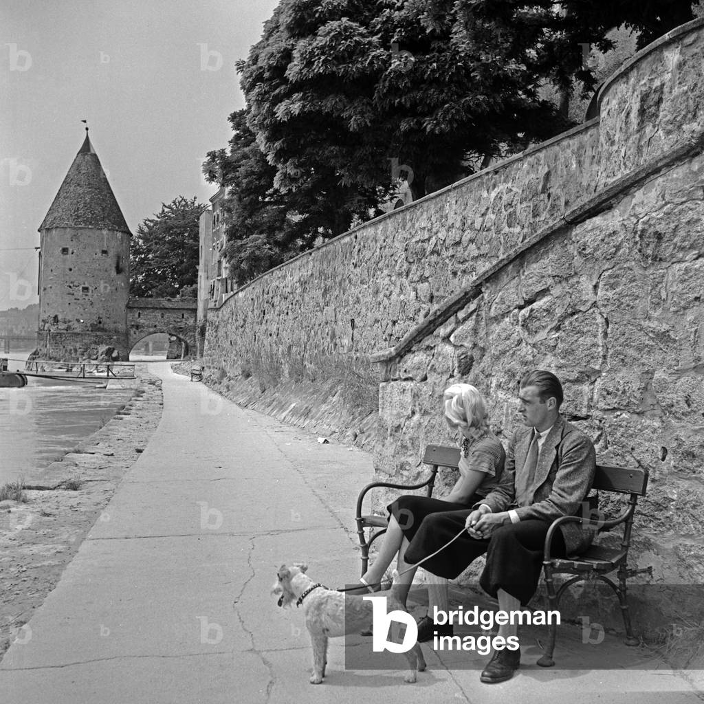 A young couple sitting on a bench at Innkai quay near Schaiblingsturm tower at Passau, Germany 1930s (b/w photo)