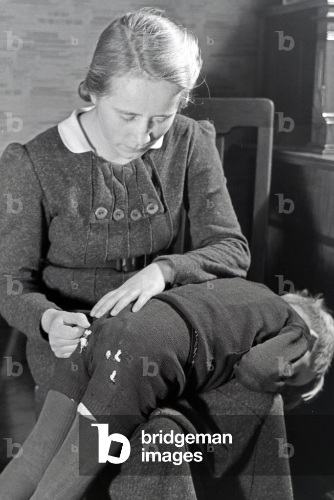 The eldest daughter of a extended family repairing the trousers of her little brother, Germany 1930s (b/w photo)