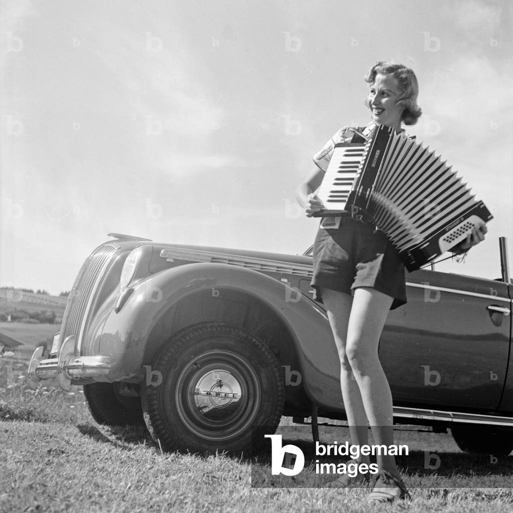 A young woman playing the accordion by an Opel model Admiral, Germany 1930s (b/w photo)
