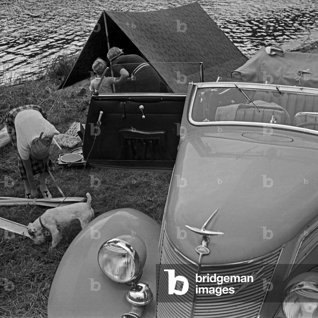 Two women packing the camping equipment into a Ford V8 after a vacation day at a lake, Germany 1930s (b/w photo)