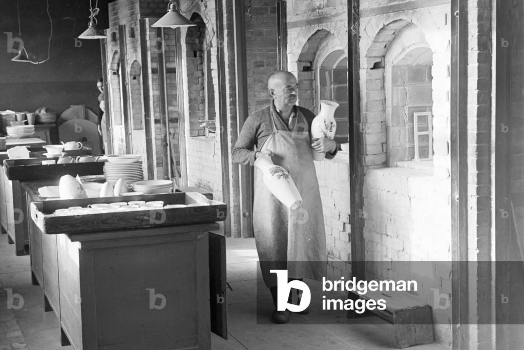 A ceramist of the State Porcelain Manufactory Berlin is carying do finished vases through the workshops, Germany 1930s (b/w photo)
