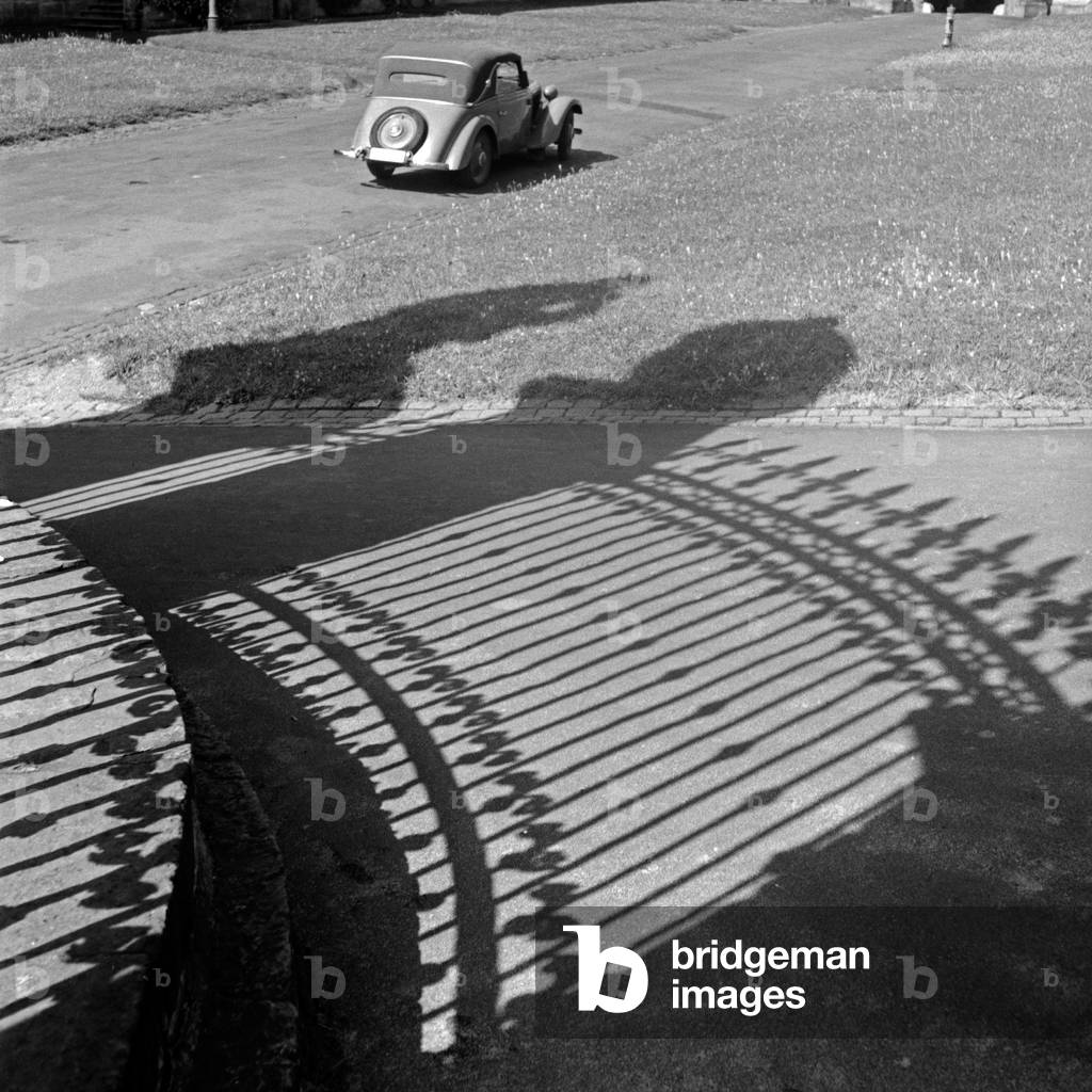 A car parking in the sun, in front of it the shadow of the wrought iron fence of Fulda orangery, Germany 1930s (b/w photo)