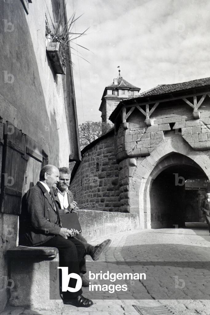 Two elderly men chitchating while sitting on a bench in front of one of the many historical archways in Rothenburg ob der Tauber, Germany 1930s (b/w photo)