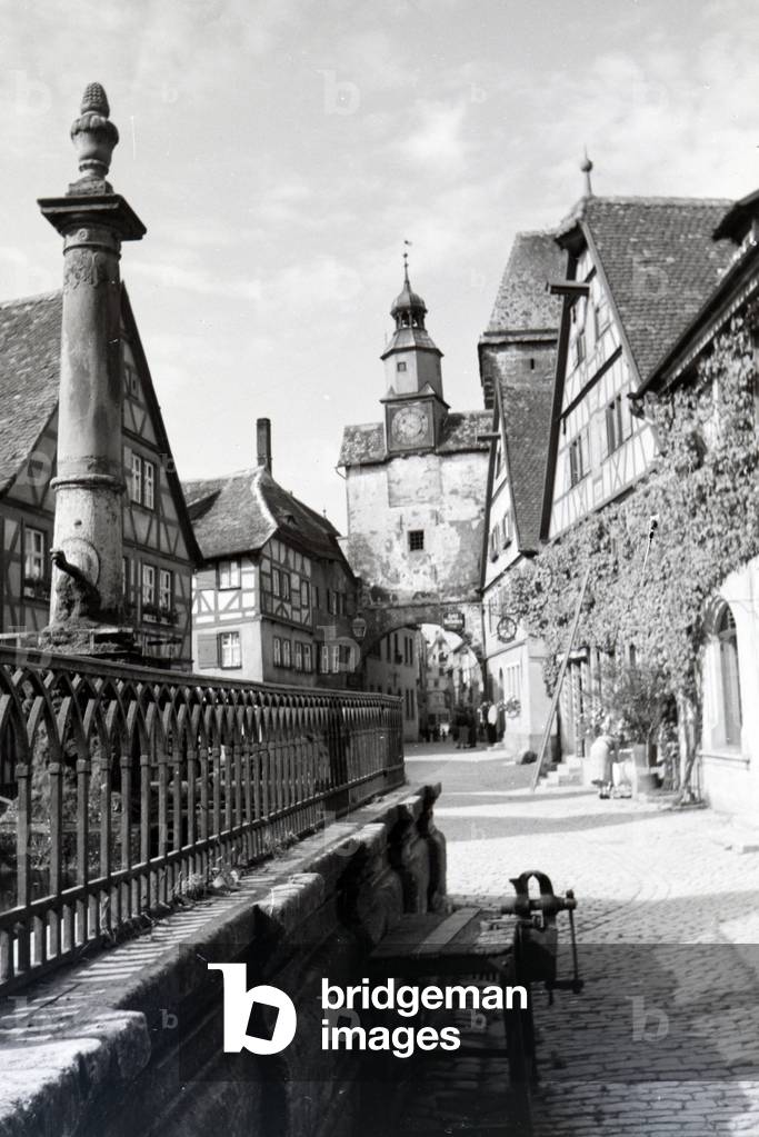 On the busy square in front of the Röder arch in Rothenburg ob der Tauber pedestrians can visit one of the many fountains of the city, Germany 1930s (b/w photo)