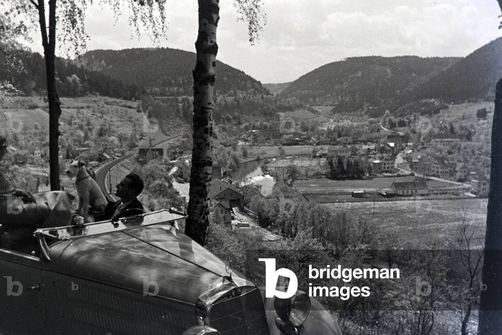 A young couple and their dog driving through Hirsau in the Northern Black Forest in a Mercedes convertible, Germany 1930s (b/w photo)