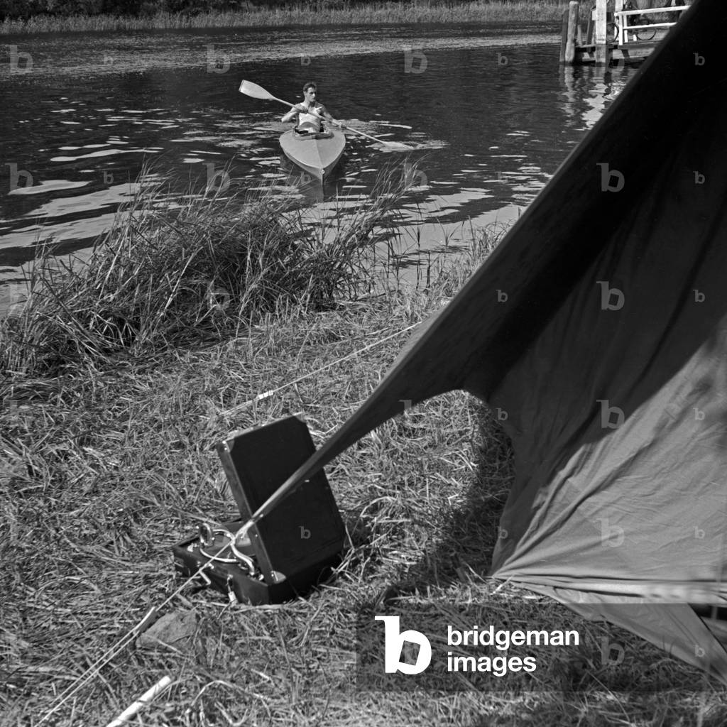 A man paddling in his Klepper folding boat to the shore of a lake, where a Klepper tent is built, Germany 1930s (b/w photo)
